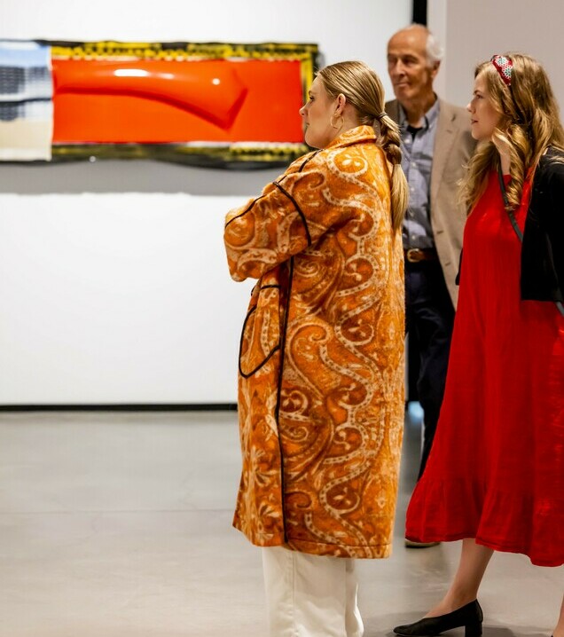 Three people stand in an art gallery, looking at a modern artwork featuring a red, abstract shape. The woman in the foreground wears an orange paisley-patterned coat and platform sandals, while another woman in a red dress and a man in a beige blazer stand nearby, observing the art.