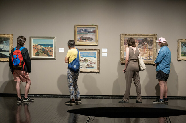 Four individuals standing in an art gallery, observing various framed paintings of landscapes hung on a dark wall.