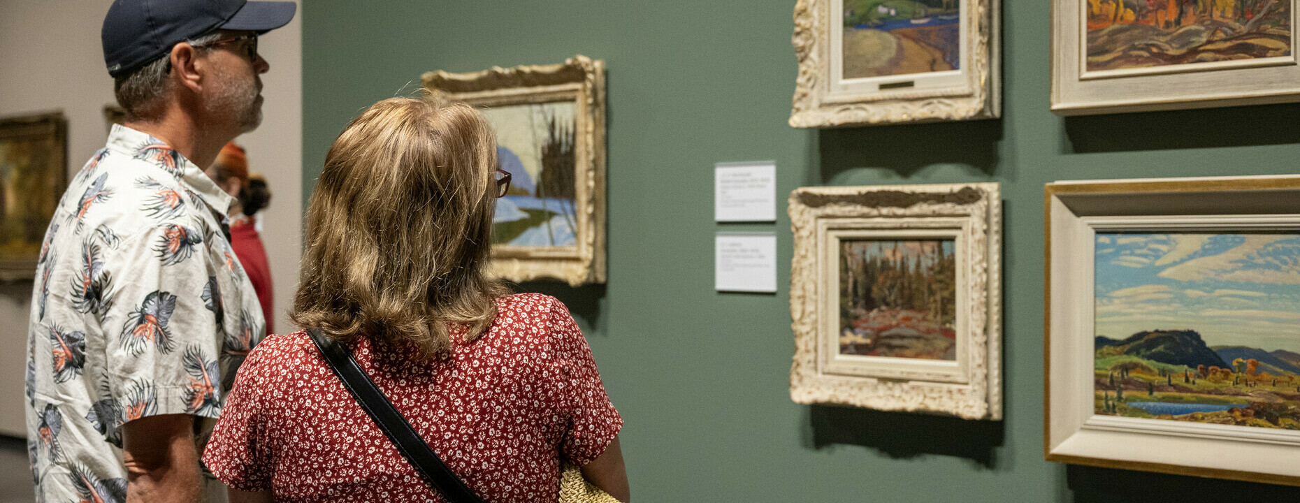 A group of people explore framed landscape paintings on a tan wall.