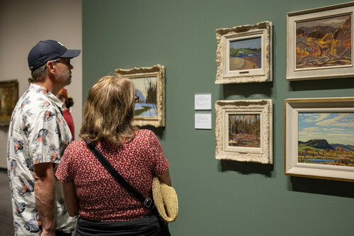 A group of people explore framed landscape paintings on a tan wall.