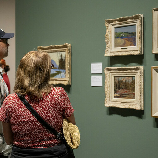 A group of people explore framed landscape paintings on a tan wall.