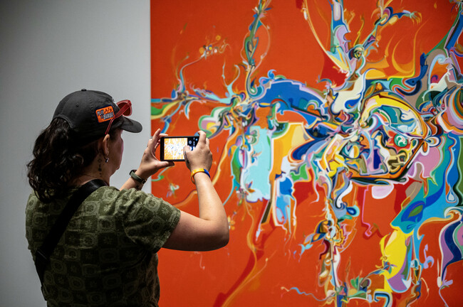 A woman takes a picture of a bright red canvas splattered with multicoloured paint.