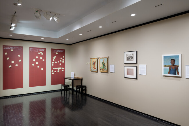 This image shows an art gallery interior with beige walls and a dark tiled floor. On the left side, there are three large red panels with white text and graphics, and a small wooden bench against the wall. On the right side, there are four framed pieces of artwork, including two paintings, one photograph, and one piece of text or document, each with a small descriptive plaque below it.