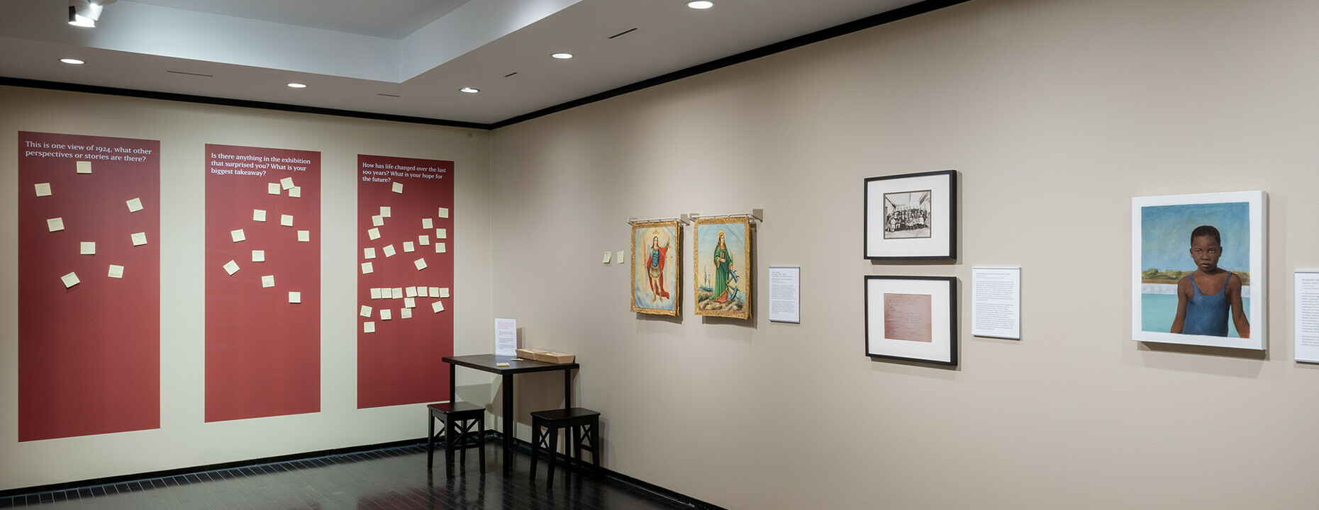 The image shows an art gallery interior with beige walls and a dark tiled floor. On the left wall, there are three large red panels with white text and graphics, followed by a small wooden bench and a music stand with some sheets. The right wall features four framed pieces of artwork, each accompanied by a small descriptive plaque