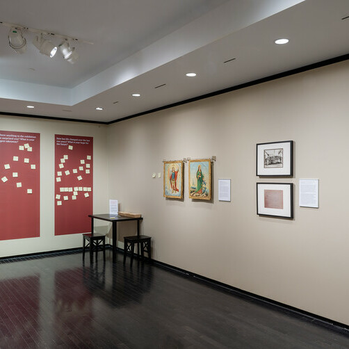 The image shows an art gallery interior with beige walls and a dark tiled floor. On the left wall, there are three large red panels with white text and graphics, followed by a small wooden bench and a music stand with some sheets. The right wall features four framed pieces of artwork, each accompanied by a small descriptive plaque