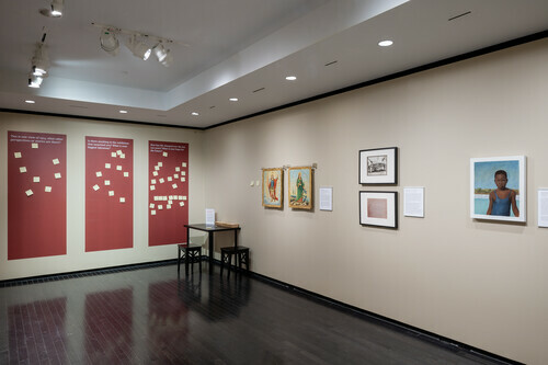 The image shows an art gallery interior with beige walls and a dark tiled floor. On the left wall, there are three large red panels with white text and graphics, followed by a small wooden bench and a music stand with some sheets. The right wall features four framed pieces of artwork, each accompanied by a small descriptive plaque