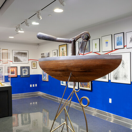 An art gallery with white walls and a polished floor, featuring an exhibition of various artworks. In the foreground, there is a large wooden sculpture resembling a boat or vessel with a metal handle attached on top, mounted on a metal stand. The background displays numerous framed artworks of different sizes and styles hung against the blue-painted sections of the wall.