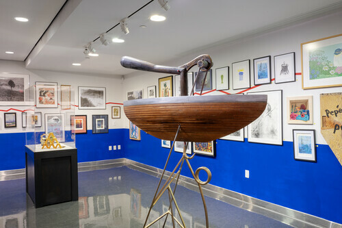 An art gallery with white walls and a polished floor, featuring an exhibition of various artworks. In the foreground, there is a large wooden sculpture resembling a boat or vessel with a metal handle attached on top, mounted on a metal stand. The background displays numerous framed artworks of different sizes and styles hung against the blue-painted sections of the wall.