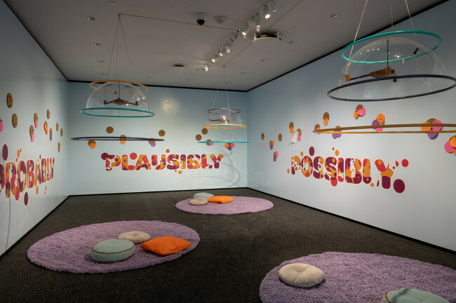 This image shows an art installation featuring a room with light blue walls and a gray carpet. Suspended from the ceiling are circular frames with transparent centers, each adorned with colorful shapes. The walls are decorated with the words “PROBABLY,” “PLAUSIBLY,” and “POSSIBLY” in bold, stylized fonts, surrounded by scattered colorful shapes that match those hanging from the frames.