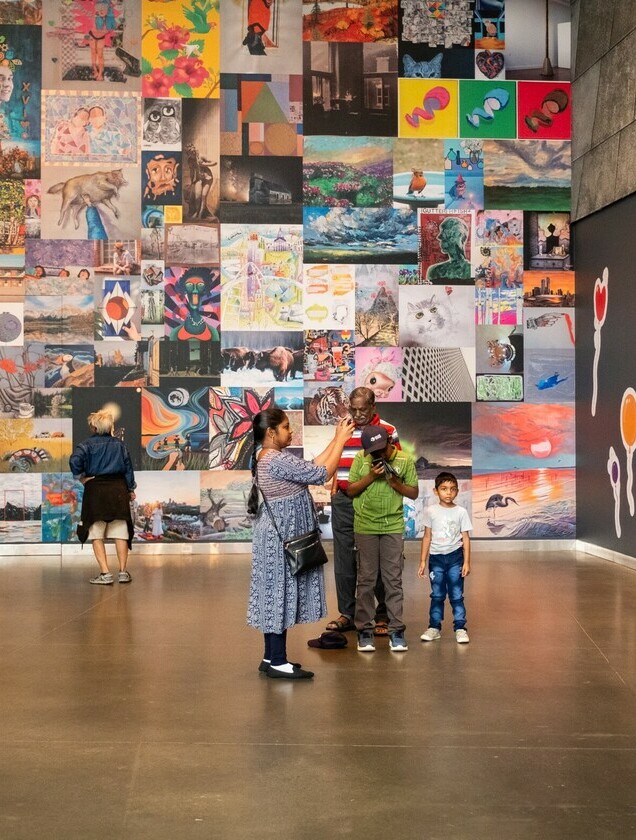 Two individuals stand in front of the AGA100 Free for All Anniversary Wall, a multicoloured wall that is filled with artwork.