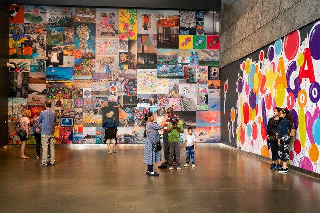 The image shows an interior view of a gallery with people observing large, colorful collages on the walls. The left wall features a grid of various images including photographs and artworks, while the right wall displays a vibrant pattern with numerous circles and dots in different sizes and colors.