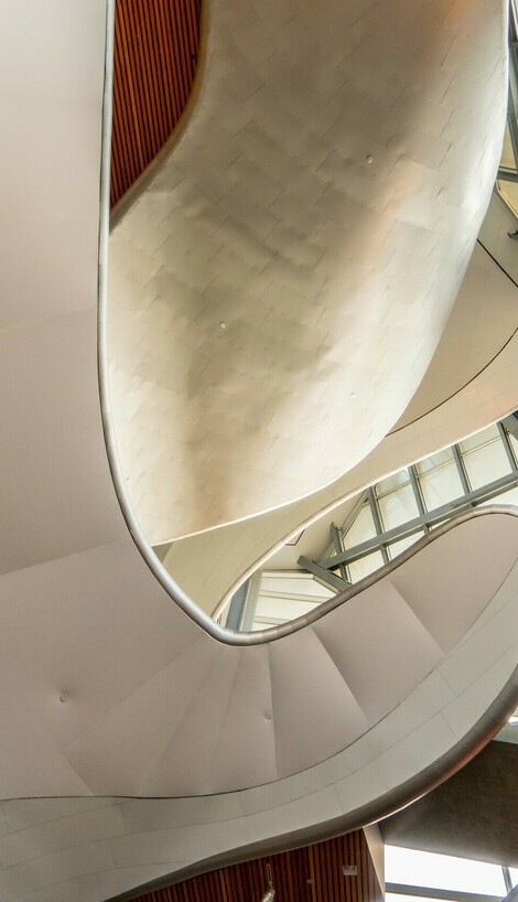 Interior view of the Art Gallery of Alberta featuring sweeping, curved architectural elements made of smooth metal and glass. The fluid, organic design contrasts with the large glass windows that allow natural light to flood the space.